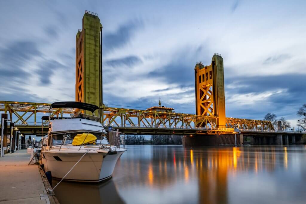 Tower Bridge in Sacramento lit up at dusk with a docked boat in the foreground and reflections on the river.